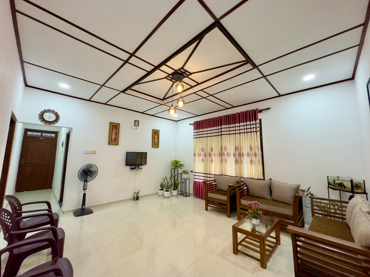 The living room features a seating area with comfortable wooden furniture set against a light-colored wall. A small coffee table is positioned in the center, complemented by potted plants. Natural light filters through the curtains, enhancing the room's brightness.