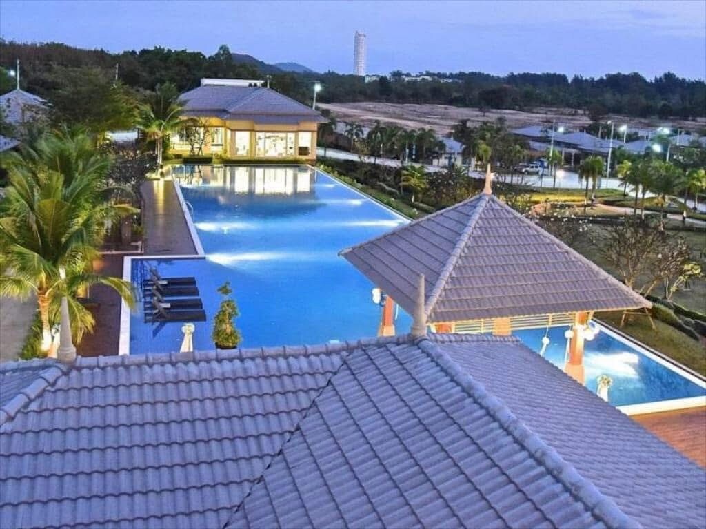 An expansive outdoor area features a large swimming pool surrounded by lush greenery and palm trees. The pool reflects evening lights, emphasizing its inviting blue water. A covered seating area is visible nearby, providing a serene spot to relax and enjoy the surroundings.