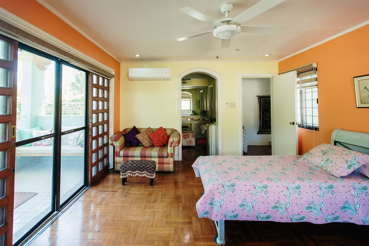 The master bedroom features a spacious bed dressed in a floral pink covering and a light-colored ceiling fan above. A cozy seating area with cushions is along one wall, while large glass doors provide access to a balcony, enhancing natural light in the room.