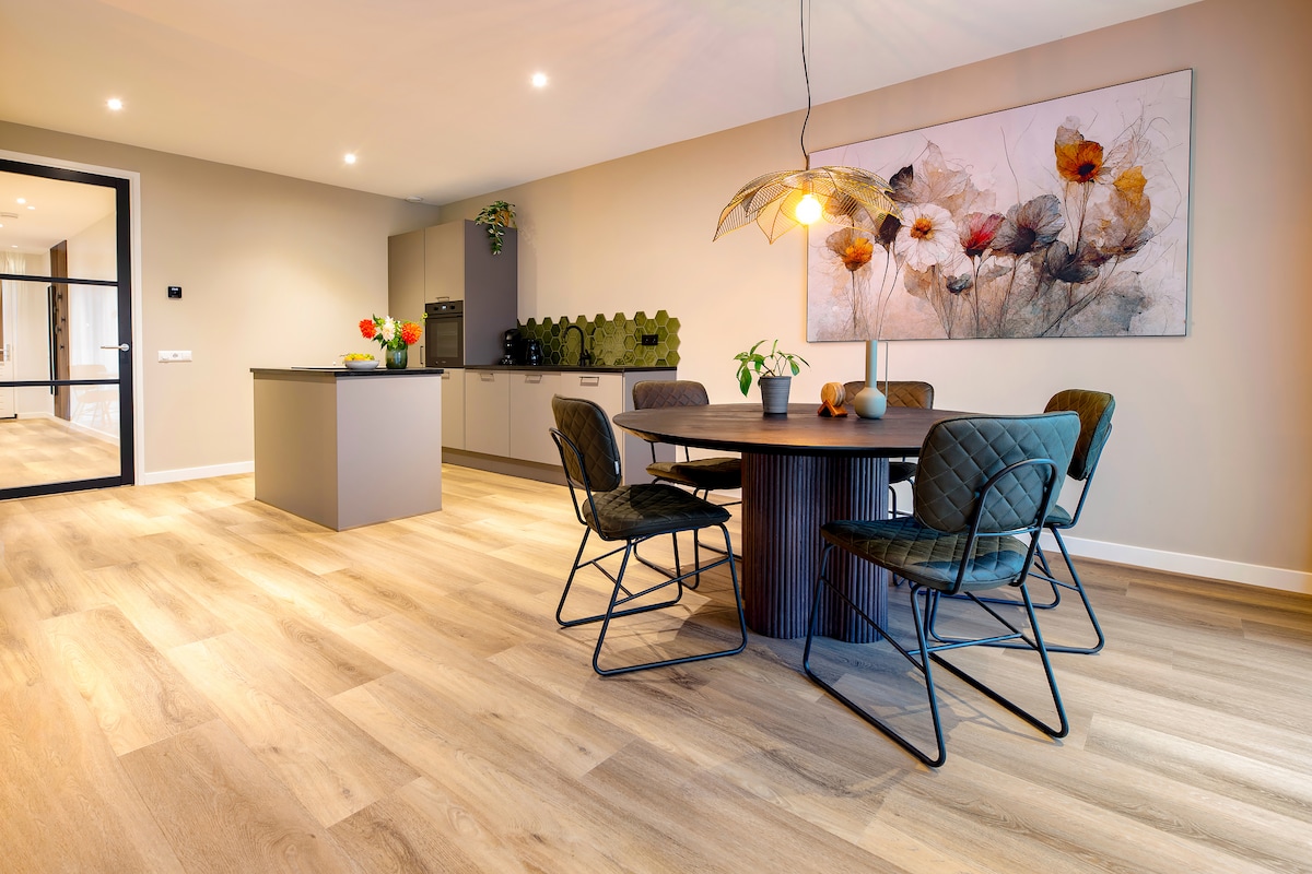 A dining area features a round table surrounded by four green chairs. A modern kitchen with a central island is visible in the background. A piece of floral artwork decorates the wall, adding a touch of color to the neutral tones of the space.