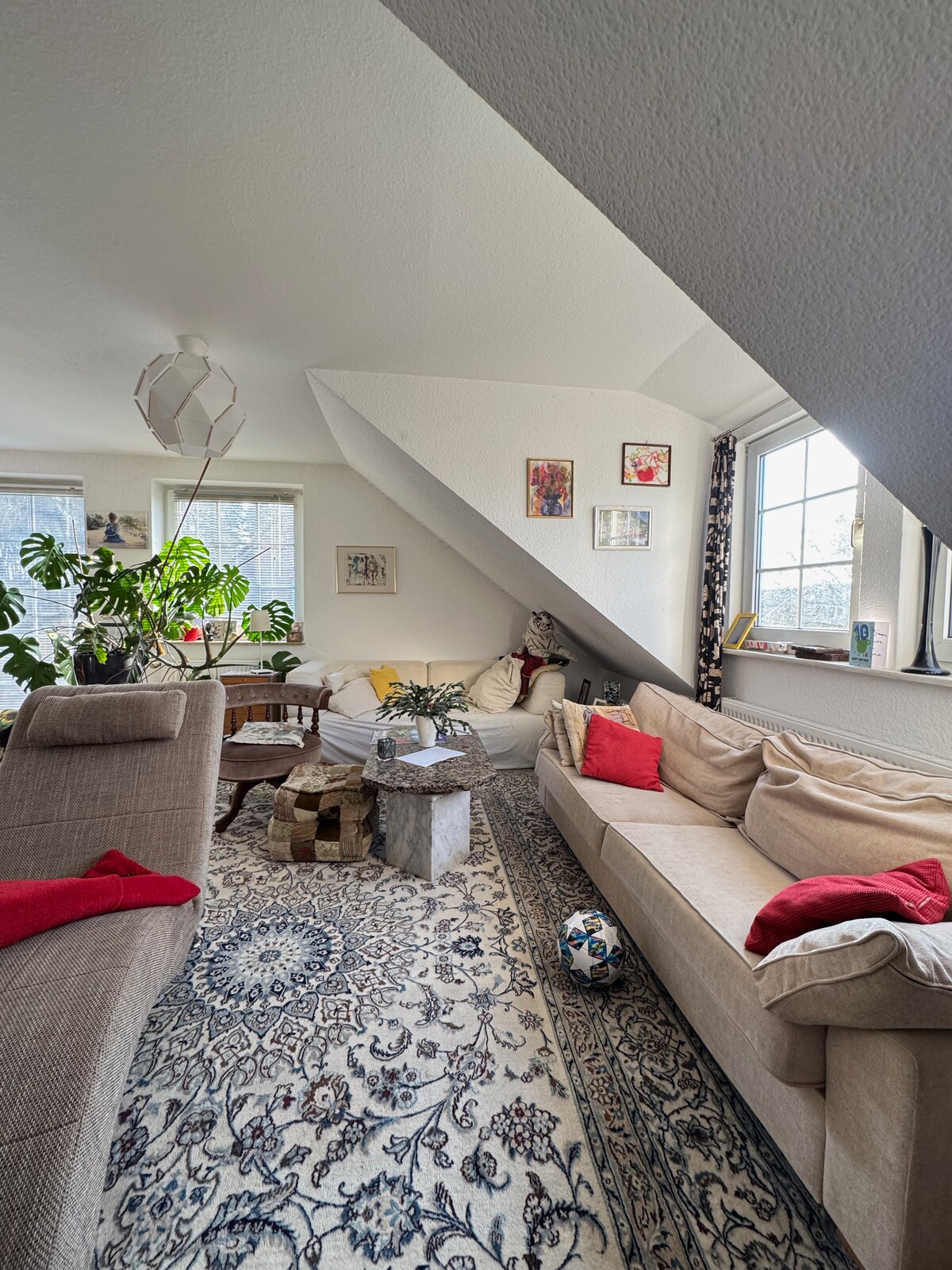A bright living room features two sofas, an armchair, and a small coffee table. A large window allows natural light to brighten the space. Decorative plants and wall art add character, while a soft area rug with intricate patterns enhances the comfort of the area.