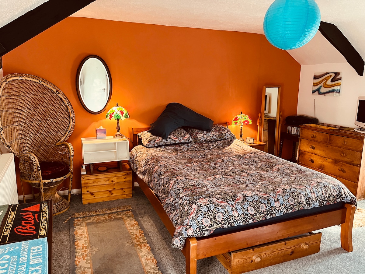 The bedroom features a spacious bed adorned with patterned bedding. Colorful lamps flank the bed, providing warm light. A round mirror is mounted on the orange wall, and a wicker chair is positioned nearby. A wooden dresser and compact TV are visible against the wall.