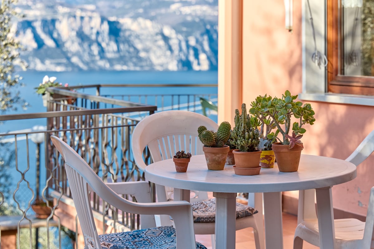 A balcony terrace is shown with a round white table surrounded by two white chairs. Potted succulent plants are arranged on the table, offering a touch of greenery against the soft peach-colored wall and providing a scenic view of the lake and mountains in the background.
