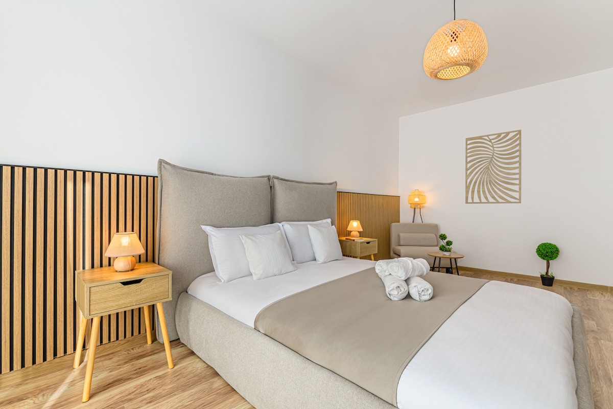 A spacious bedroom is featured with a large bed adorned with neutral-toned linens and decorative throw pillows. Table lamps on either side provide soft lighting. A cozy seating area with an accent chair is visible, complemented by a wall decoration and a small plant.