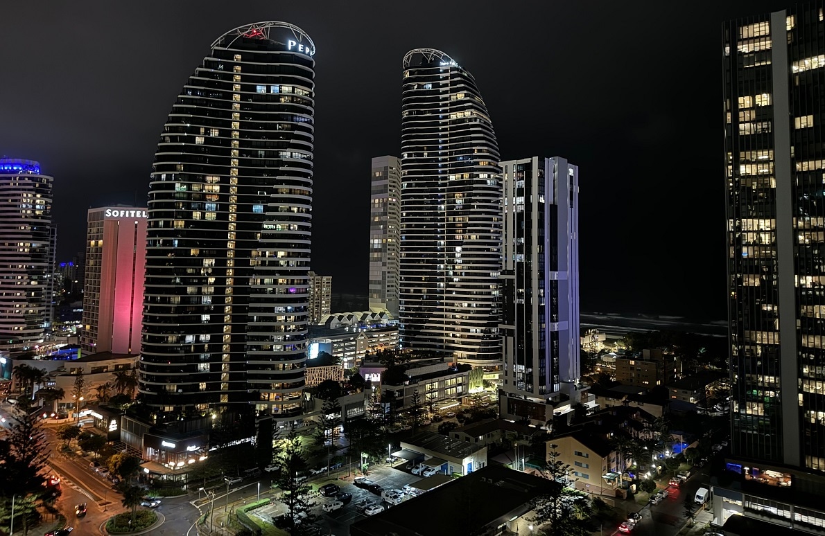 A night view showcases modern high-rise buildings illuminated by vibrant lights. The ocean is visible in the background, while a mix of busy streets and city life unfolds below. The varied architectural styles contribute to the dynamic skyline.
