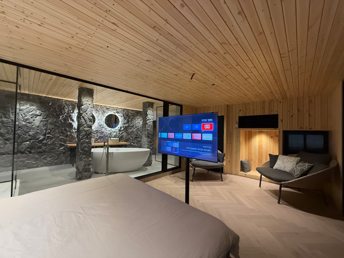 A modern bedroom features a spacious bed with a light-colored duvet. A large TV screen displays various media options. The room is bordered by glass walls, providing a view of a stylish bathroom with a freestanding tub and unique stone accents.