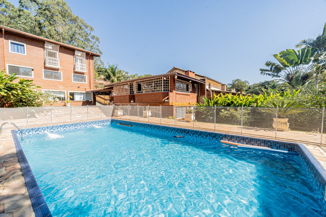 A large outdoor pool invites relaxation, surrounded by a fence for safety. Lush greenery frames the area, with a well-maintained brick structure visible in the background. Clear blue skies create a serene atmosphere, enhancing the inviting setting for leisure activities.