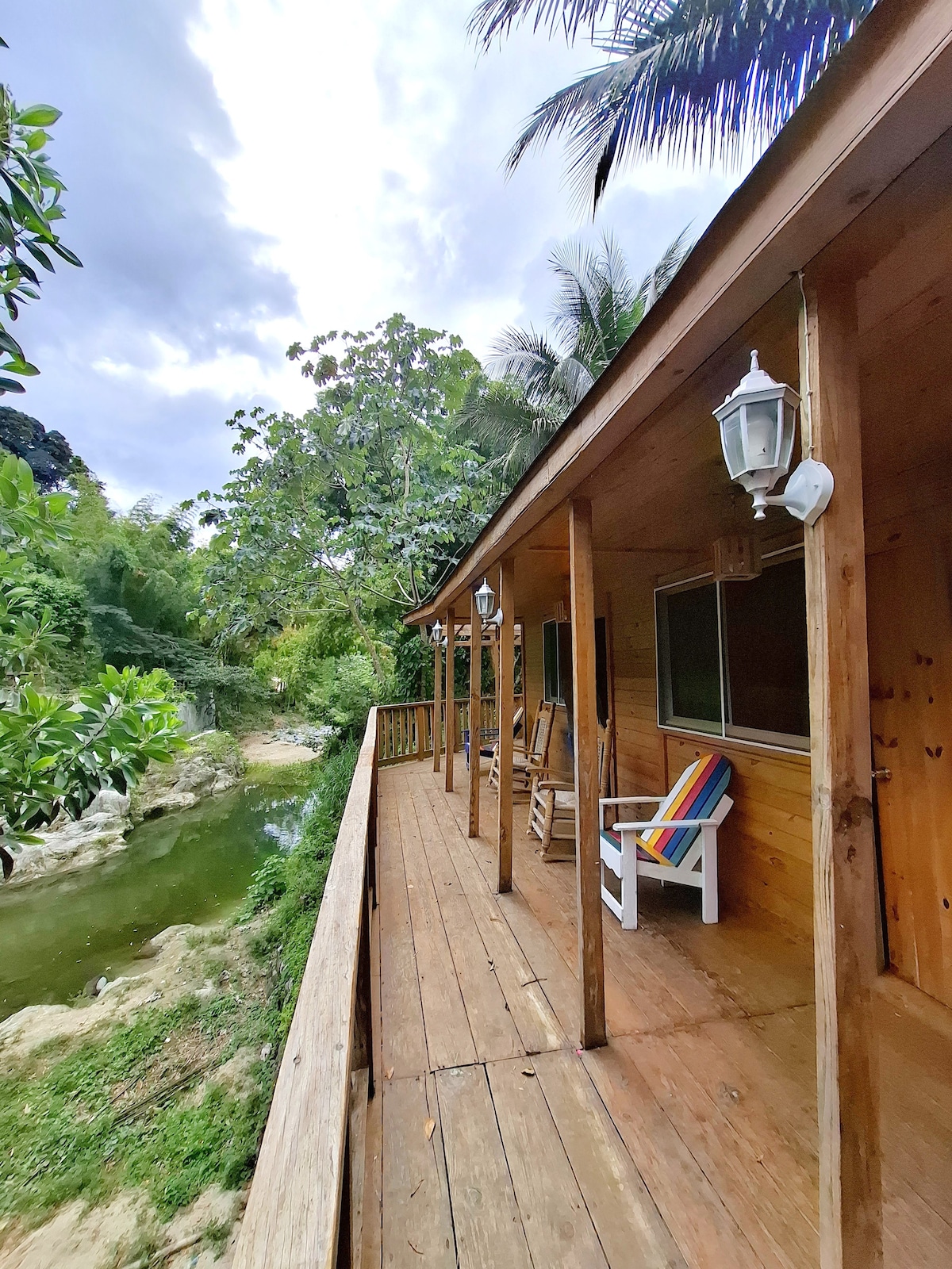 A wooden deck extends alongside the cabin, featuring colorful chairs, inviting relaxation. Lush greenery surrounds the area, with the Mahomita River visible nearby. Overhead lanterns provide subtle illumination, enhancing the natural setting. The cabin's exterior showcases a rustic charm in a tranquil environment.