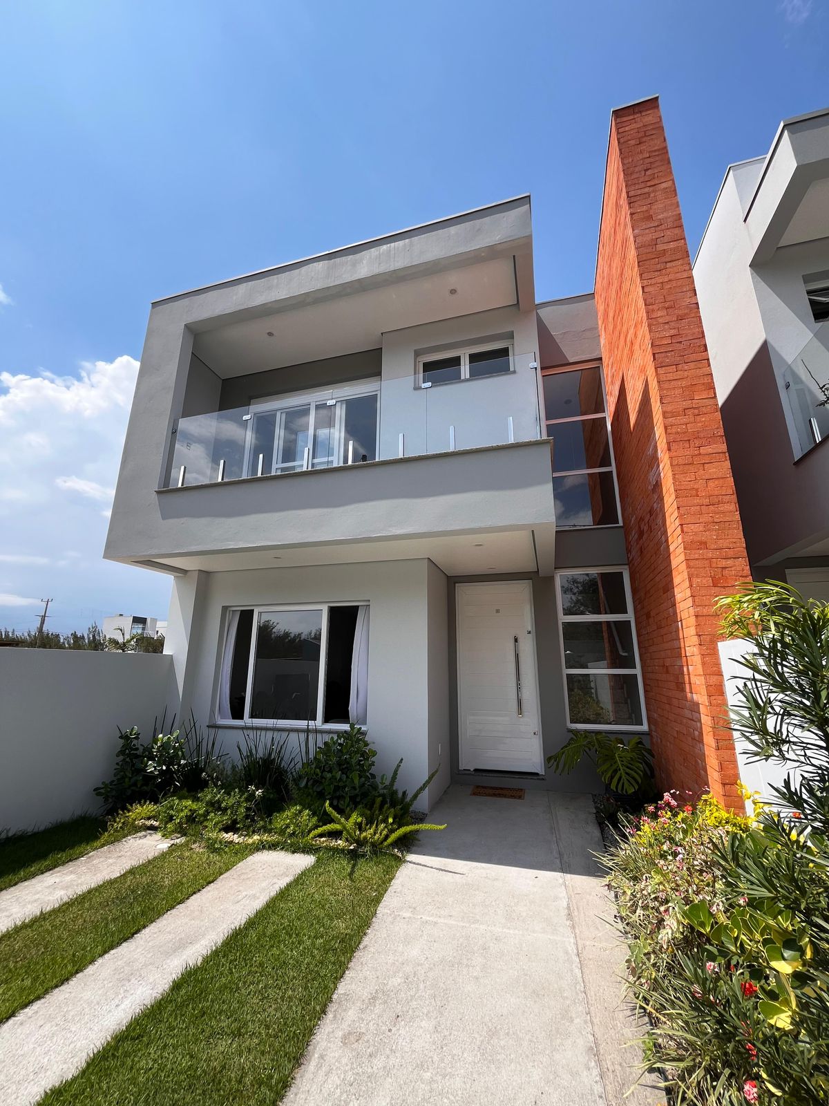 The exterior of the house features a modern design with two stories, highlighted by a combination of light and reddish-orange finishes. A welcoming entrance is flanked by well-kept greenery and landscaping, with a concrete pathway leading to the front door.