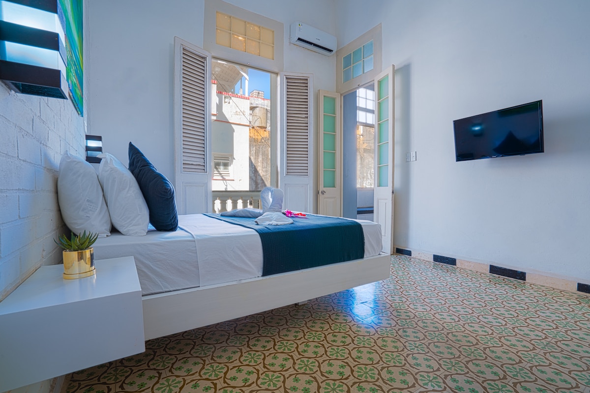 A well-lit bedroom features a spacious bed with crisp white linens and a navy blue accent. Large windows with wooden shutters allow natural light to fill the room. A wall-mounted television is visible, and a small plant adds a touch of greenery.