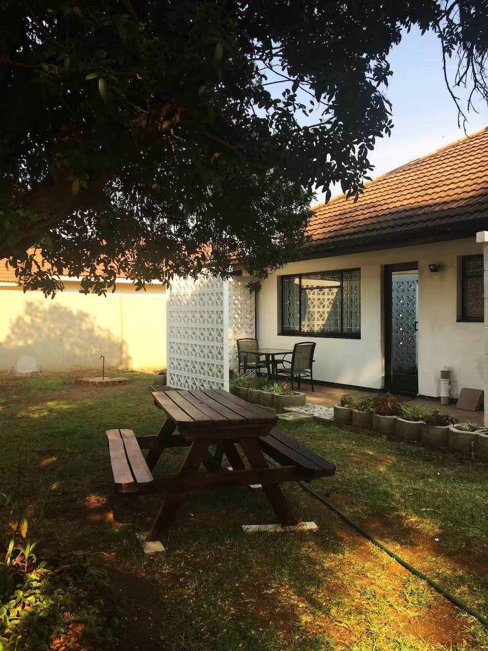 A shaded outdoor area features a wooden picnic table surrounded by grass. Nearby is a small patio with a black metal table and chairs. A white lattice wall adds privacy, and a tree provides additional shade to the space.