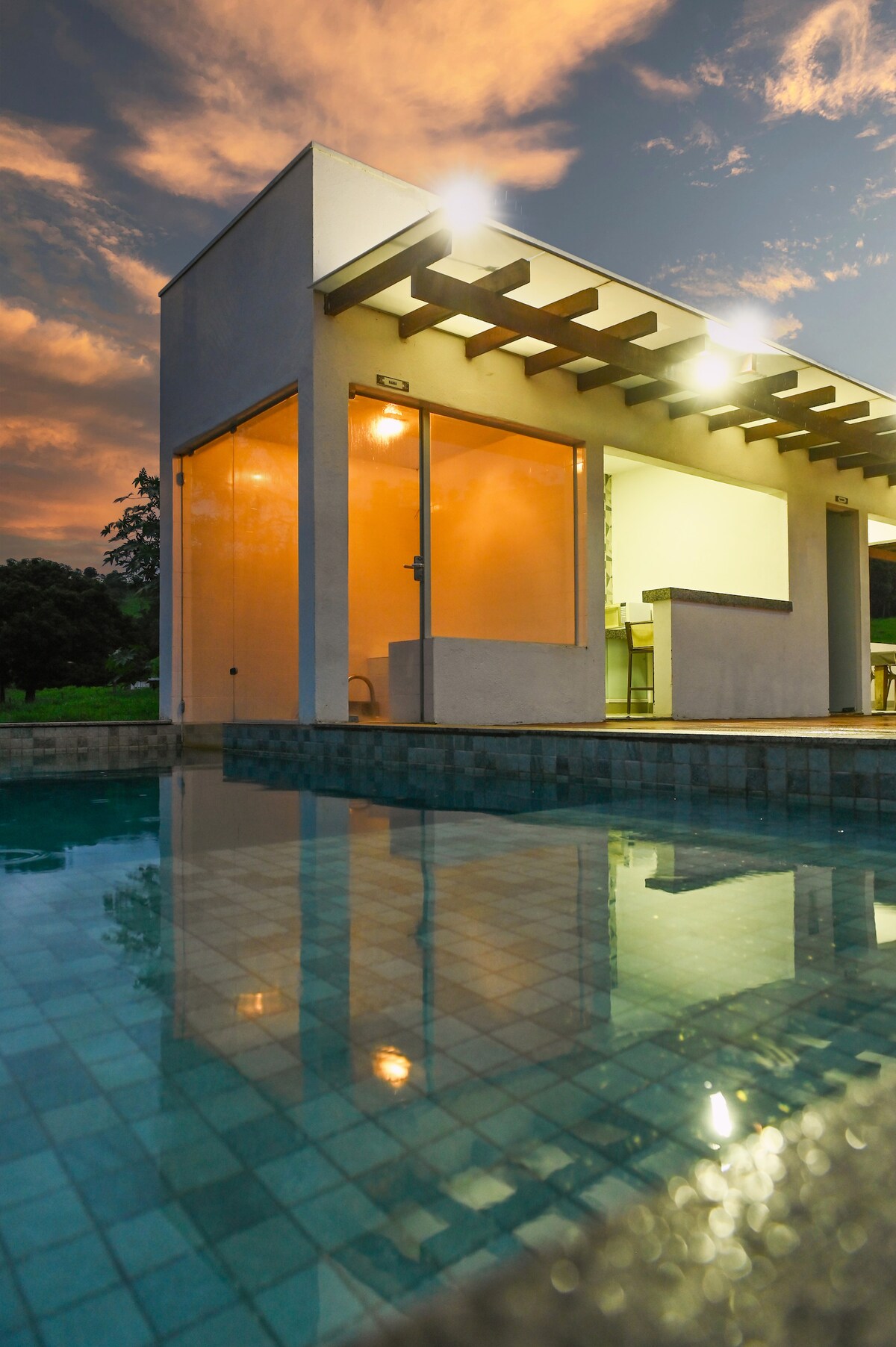 A modern structure with large glass walls is reflected in the calm waters of a swimming pool. The soft lighting from the interior casts a warm glow against the evening sky, which features hues of orange and pink among scattered clouds.