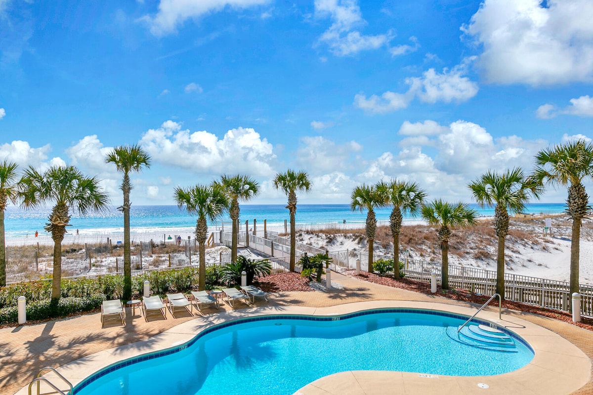 A swimming pool is surrounded by palm trees, with lounge chairs arranged nearby. In the background, the beach and ocean are visible under a bright blue sky dotted with fluffy clouds. A pathway leads from the pool area to the sandy shore.
