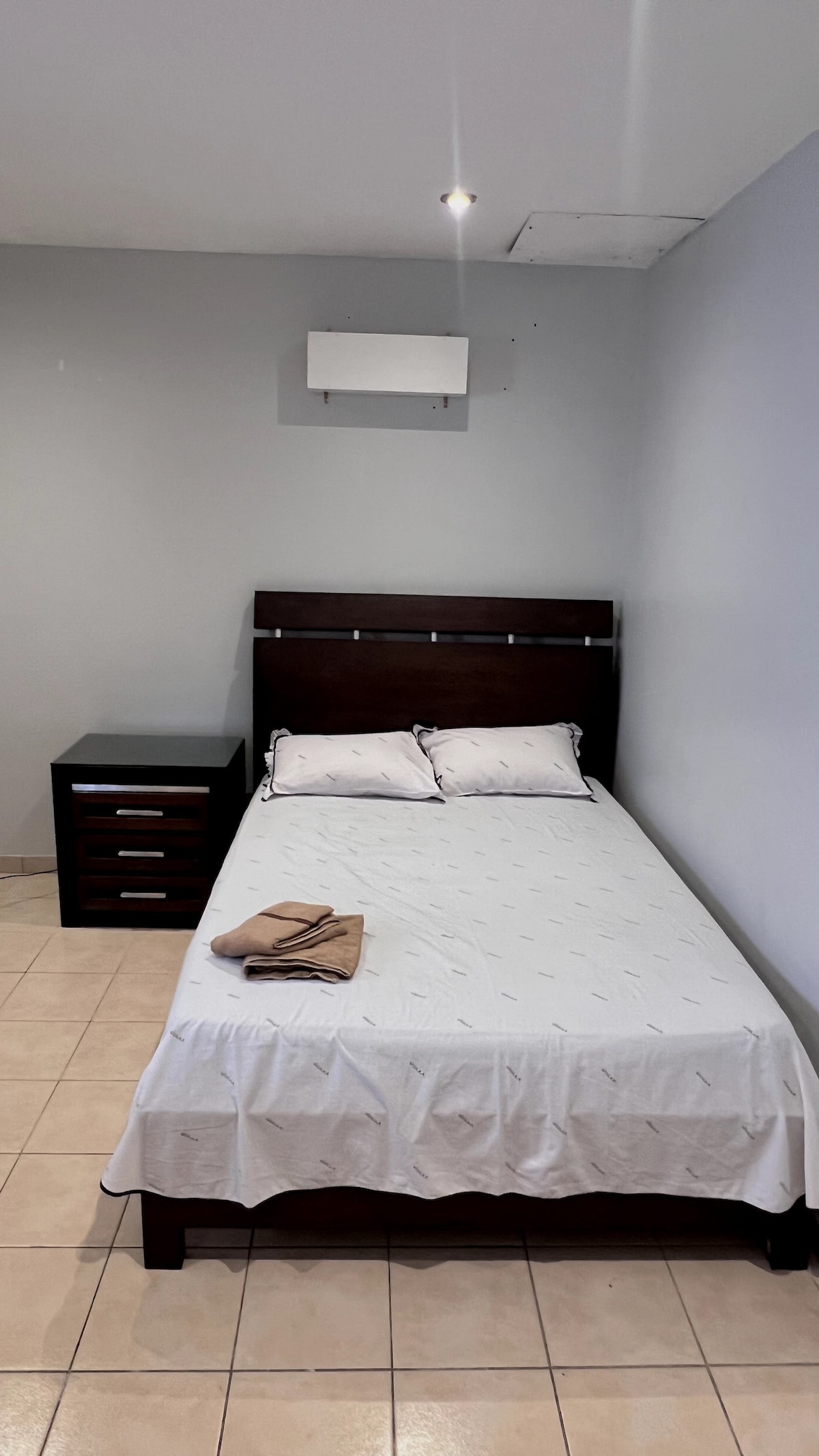 A well-lit room features a single bed with a neatly arranged white bedspread and two pillows. A dark wood nightstand with drawers sits beside the bed. Light tiles cover the floor, and a wall-mounted air conditioning unit is positioned above the bed.