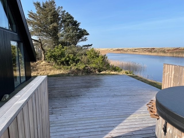 A spacious wooden terrace is shown overlooking a calm waterway, framed by lush green shrubs and tall grass. The structure features a hot tub, with clear skies visible in the background, creating a serene outdoor space for relaxation.