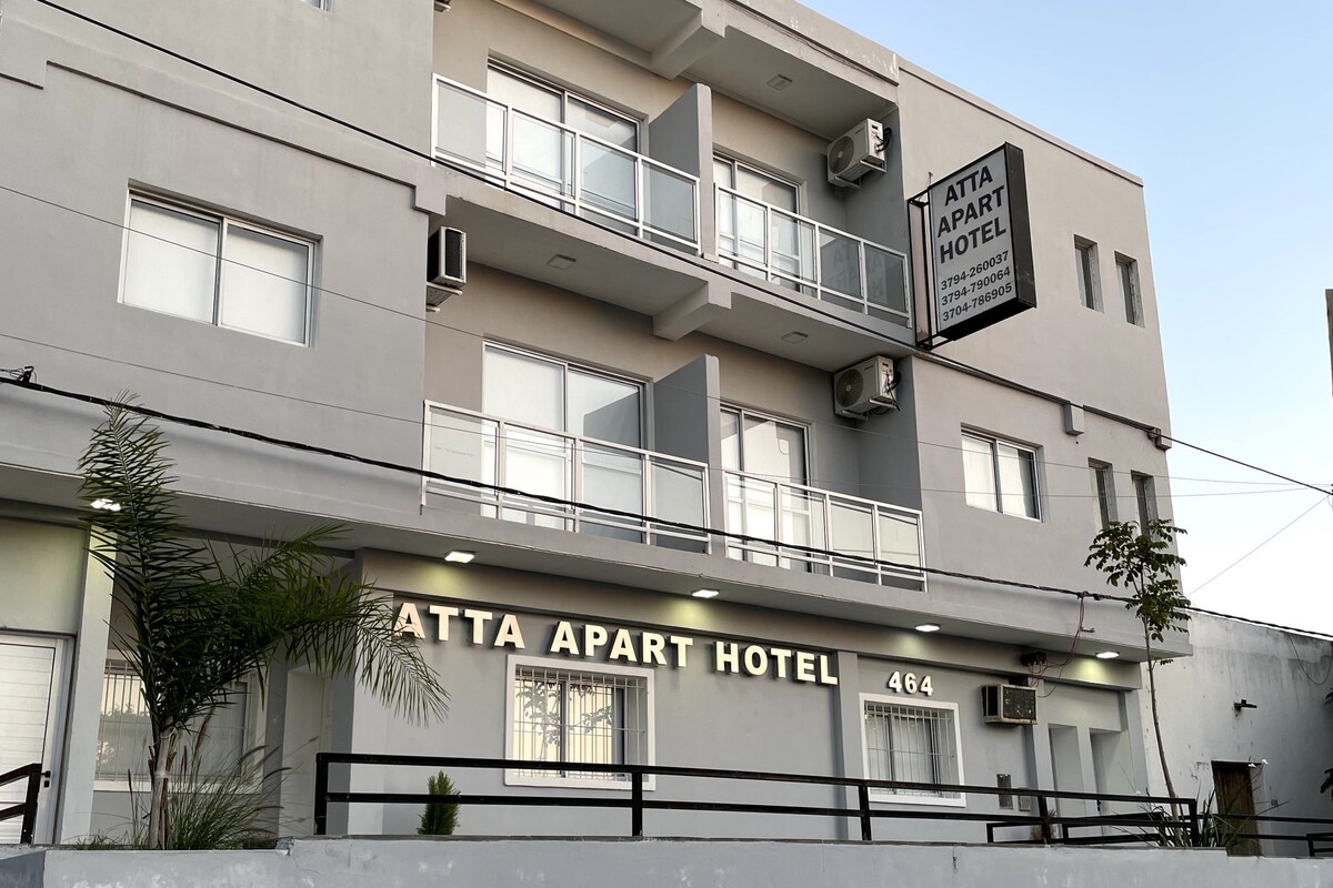 The exterior of a modern building houses ATT Apart Hotel, featuring a neutral color palette and multiple balconies. The ground-level entrance is marked by a clear sign displaying the hotel name and street number. Potted plants add a touch of greenery to the facade.