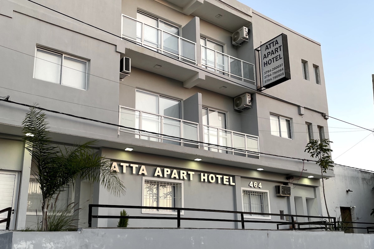 A modern building with a light gray facade is shown, featuring multiple balconies and large windows. The hotel name is clearly displayed above a well-lit entrance. Lush greenery along the pathway adds a touch of nature to the urban setting.