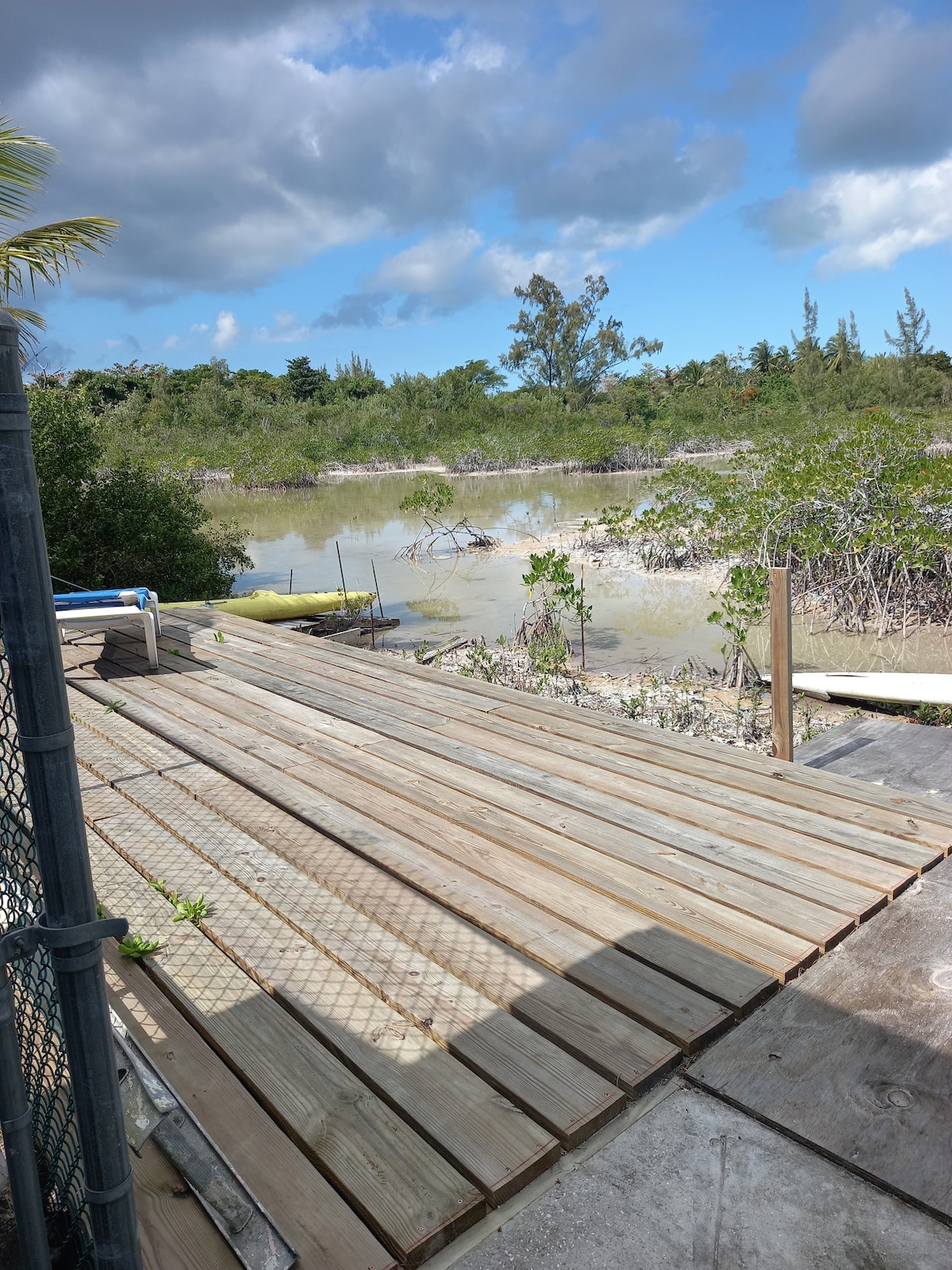 A wooden deck extends towards a natural landscape, featuring mangroves and shallow waters. The area is surrounded by lush greenery, offering a peaceful view. The sky above is partly cloudy, contributing to a serene atmosphere.
