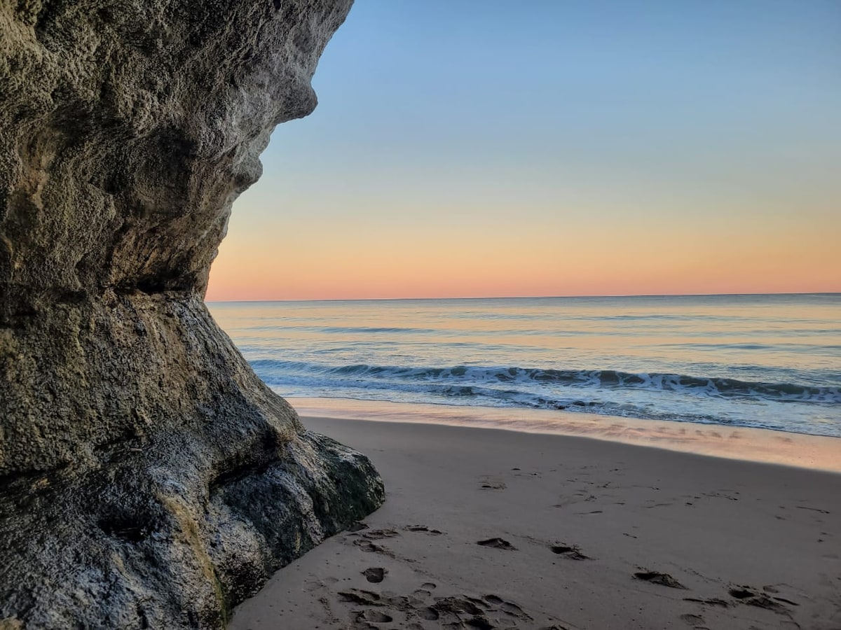 A serene beach scene is captured, with soft waves lapping against the shore. A rocky formation frames the view on the left, contrasting against a pastel sky that transitions from warm hues to cooler tones. Traces of footprints are visible in the sandy foreground.