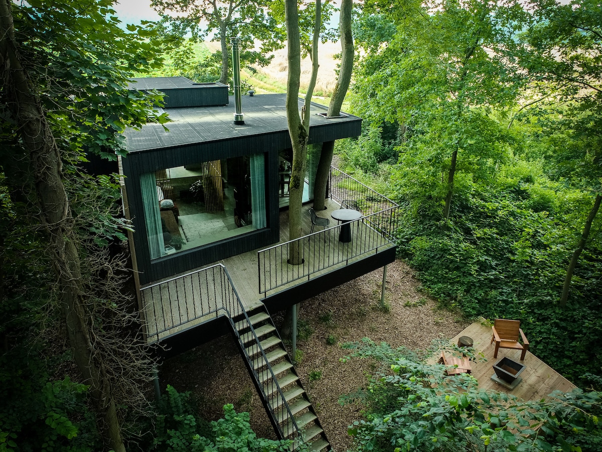 A modern structure sits elevated among trees, featuring large glass windows that reveal the interior. A deck is accessible via a staircase, with a small outdoor area furnished with a single chair and a fire pit, surrounded by lush greenery.