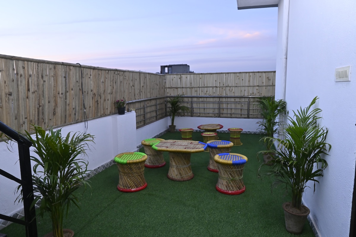 An outdoor seating area features multiple round tables surrounded by colorful stools. Lush green plants are placed in pots, adding freshness to the space. The area is enclosed by a wooden fence, providing a private setting with a grassy surface, ideal for relaxation.