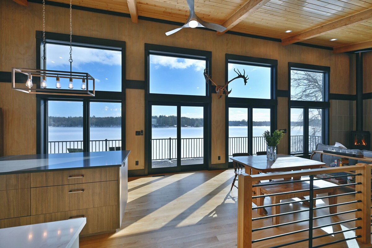 A spacious living area is illuminated by natural light from large, floor-to-ceiling windows. The view overlooks the serene waters of Horseshoe Lake, while a modern kitchen island and dining table provide ample space for family gatherings. Rustic accents enhance the inviting design.