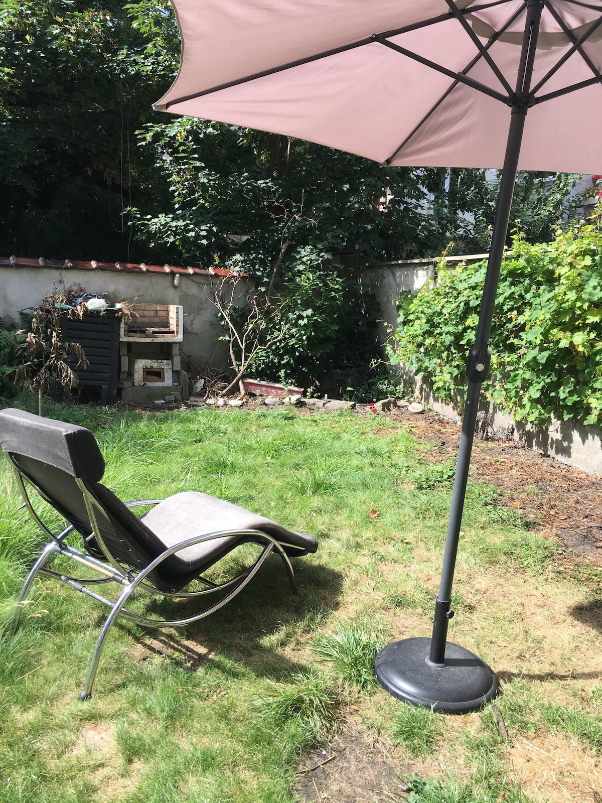 A garden area is displayed, featuring a lounge chair and a large umbrella offering shade. Lush green grass is visible, interspersed with some low shrubs and a stone wall in the background, creating a serene outdoor setting.