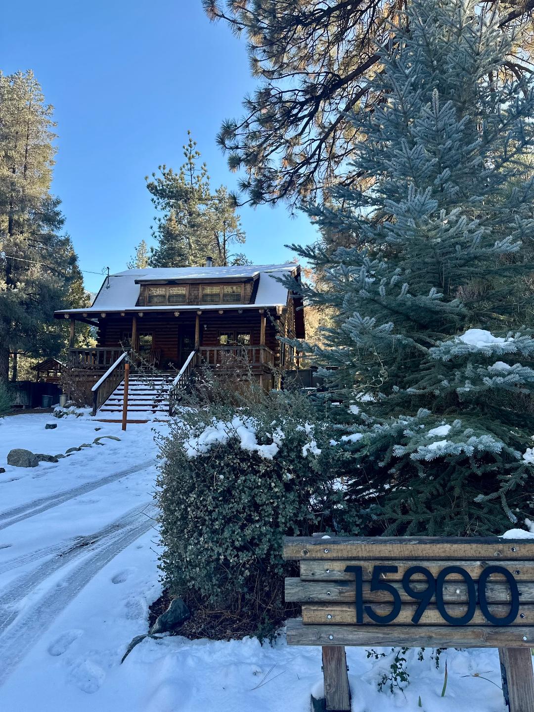 A wooden log cabin cabin is surrounded by snow-covered trees, with a sturdy staircase leading to the entrance. The property number is displayed on a wooden sign in the foreground, while the clear blue sky enhances the serene winter landscape.
