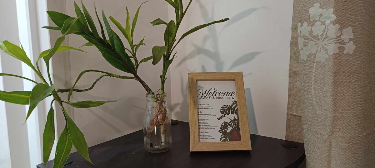 A small table holds a clear glass jar with a thriving green plant, complemented by a wooden-framed sign that reads 'Welcome.' The background features light-colored walls, enhancing the serene ambiance of the space.