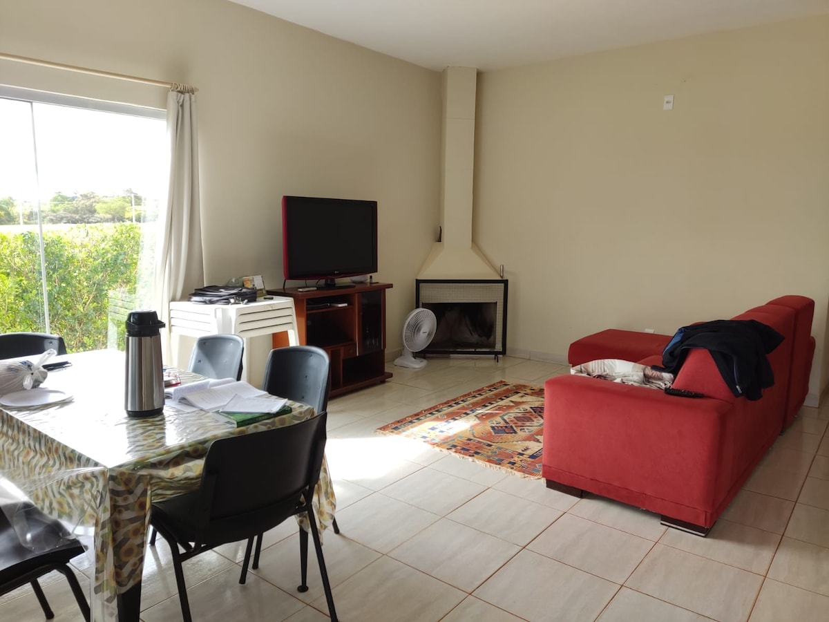 A living space is presented with a cozy red sofa facing a television. A dining table with dark chairs occupies one side, while a decorative rug adds texture to the tile floor. Natural light filters through large glass doors, enhancing the bright atmosphere.
