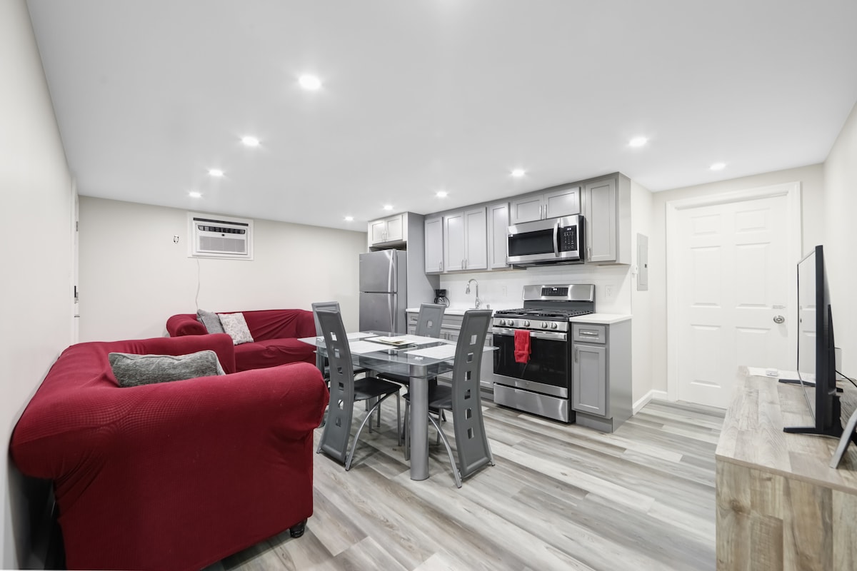 An open-concept living space features modern kitchen appliances, including a stainless steel refrigerator and oven. A dining table with gray chairs sits next to a comfortable red sofa. Recessed lighting brightens the area, highlighting the sleek laminate flooring and neutral wall colors.