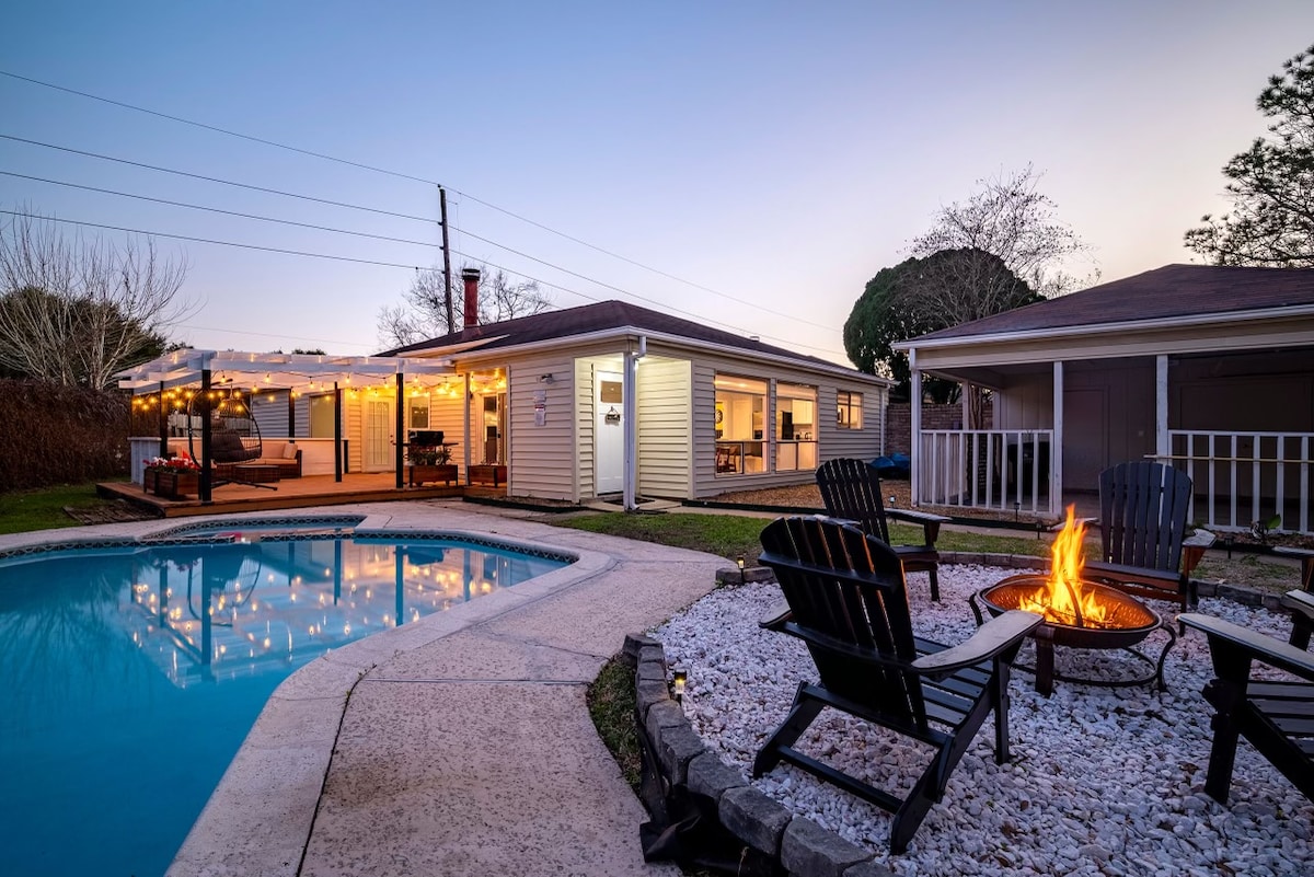 The outdoor area showcases a sparkling pool adjacent to a fire pit surrounded by black chairs, creating a cozy atmosphere. The patio is illuminated by warm ambient lighting, providing an inviting setting for gatherings. The house features a classic exterior, enhancing the serene environment.