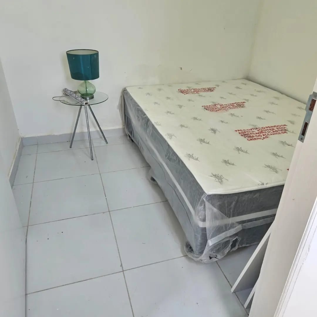 A simple bedroom is shown, featuring a spacious bed covered in a protective layer. A small round table with a green lamp is positioned beside the bed. The room presents clean, tiled flooring and white walls, creating a fresh atmosphere.