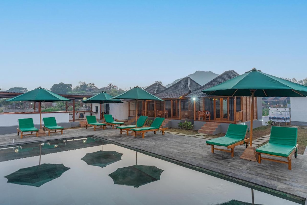 A serene outdoor area features a large swimming pool reflecting numerous green lounge chairs and umbrellas. A wooden cottage is visible in the background, with a gentle incline suggesting nearby hills. Clear skies enhance the tranquil atmosphere of the setting.