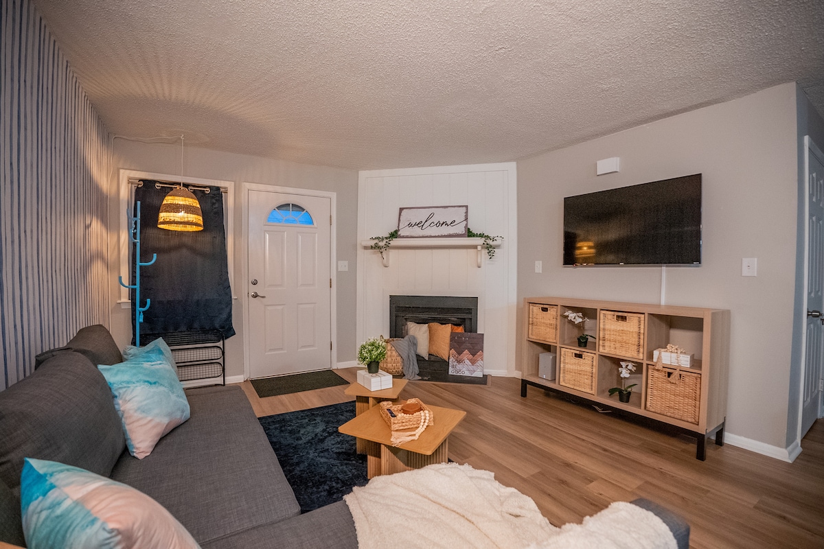 The living room features a sectional sofa adorned with decorative pillows, a cozy throw draped over a small coffee table, and a modern TV mounted on the wall. An electric fireplace, designed for decoration, is flanked by a welcoming entrance door, creating an inviting space.
