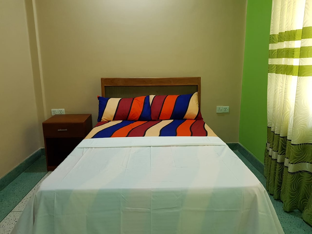 A sleeping area is presented with a spacious bed featuring a striped blanket in vibrant colors. Pillows with matching patterns are arranged neatly against the headboard. A wooden nightstand sits beside the bed, with natural light filtering through curtains adorned with subtle green stripes.