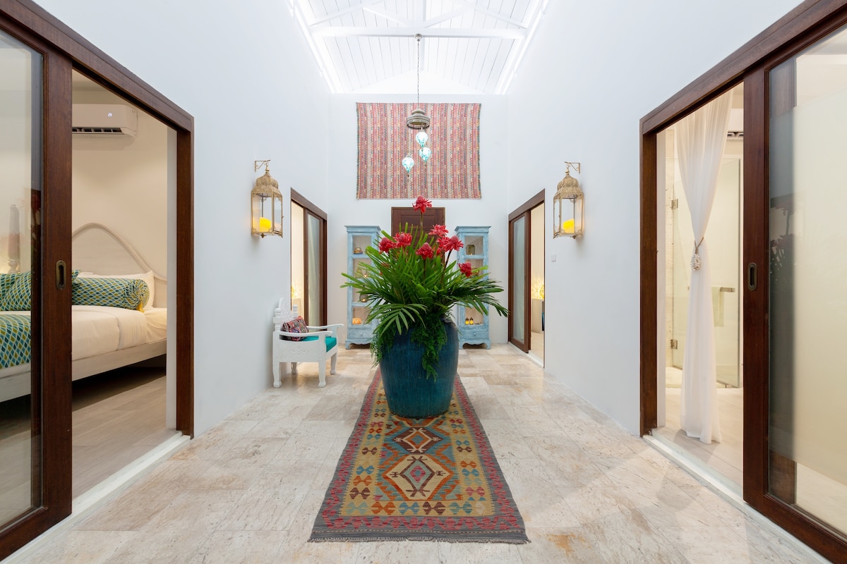 An open atrium is highlighted by a large potted plant at its center, surrounded by colorful artwork. Wooden doors lead to three separate rooms, with natural light streaming through, illuminating the stone floor and a patterned area rug that runs the length of the space.