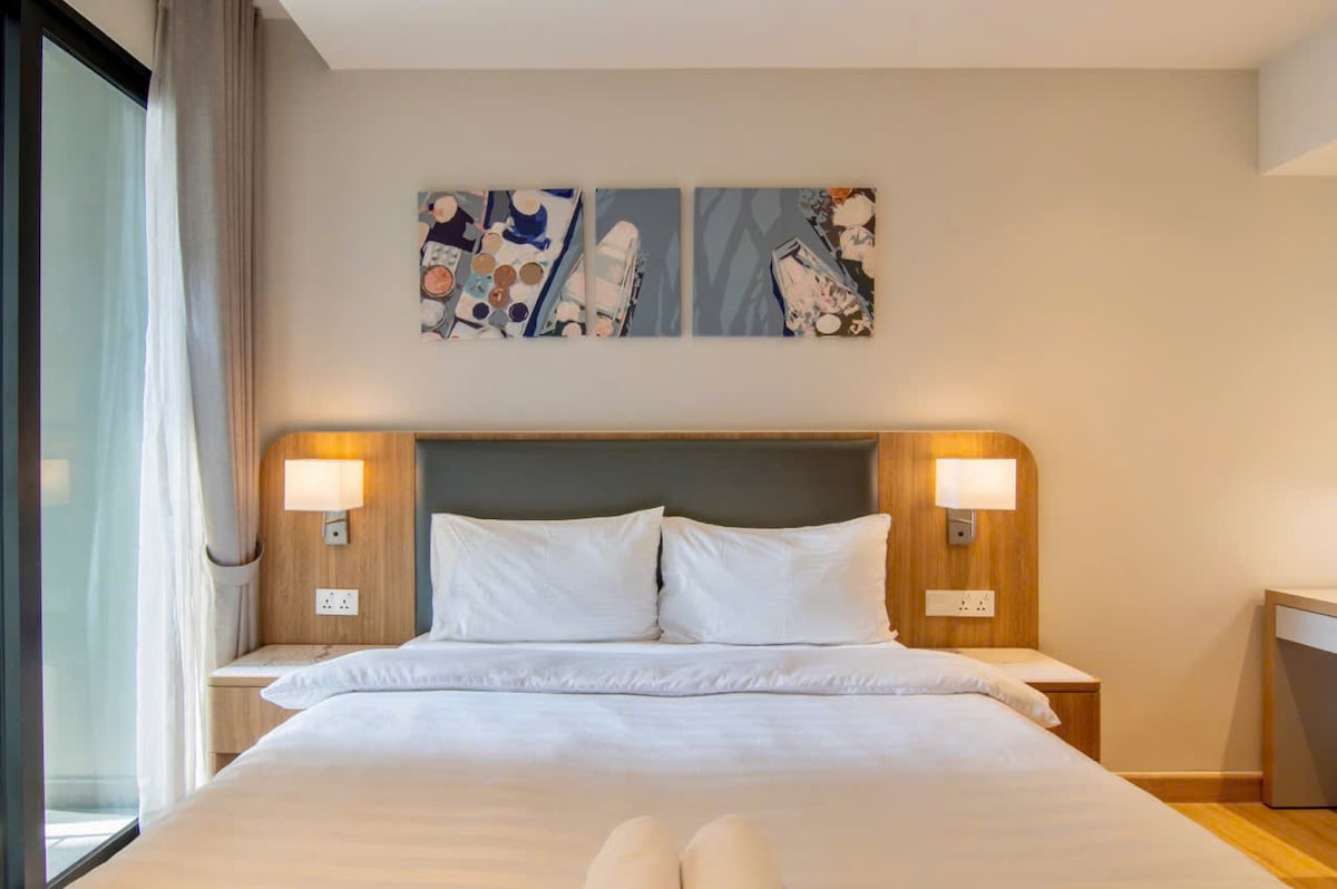 A comfortable bedroom features a spacious bed adorned with white linens and accent pillows. Above the bed, three art pieces depict serene scenes. Soft lighting is provided by bedside lamps on each side, while large windows allow natural light to fill the room.