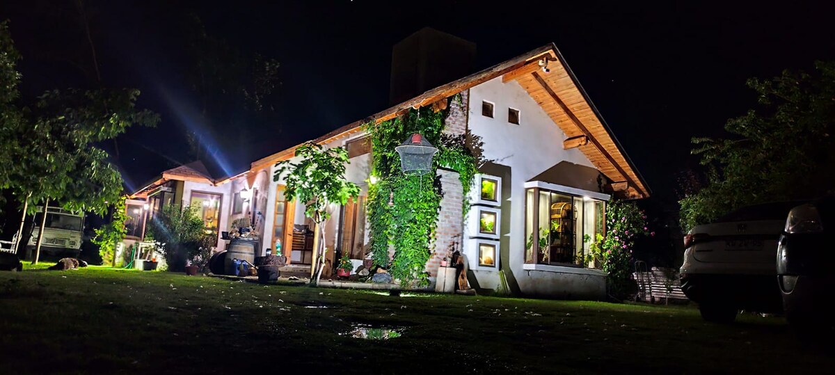 A charming exterior is illuminated at night, showcasing a stone and wood structure. Lush greenery embraces the building, while warm light spills from large windows, creating a welcoming atmosphere. The surrounding garden and driveway feature parked cars and well-maintained landscaping.