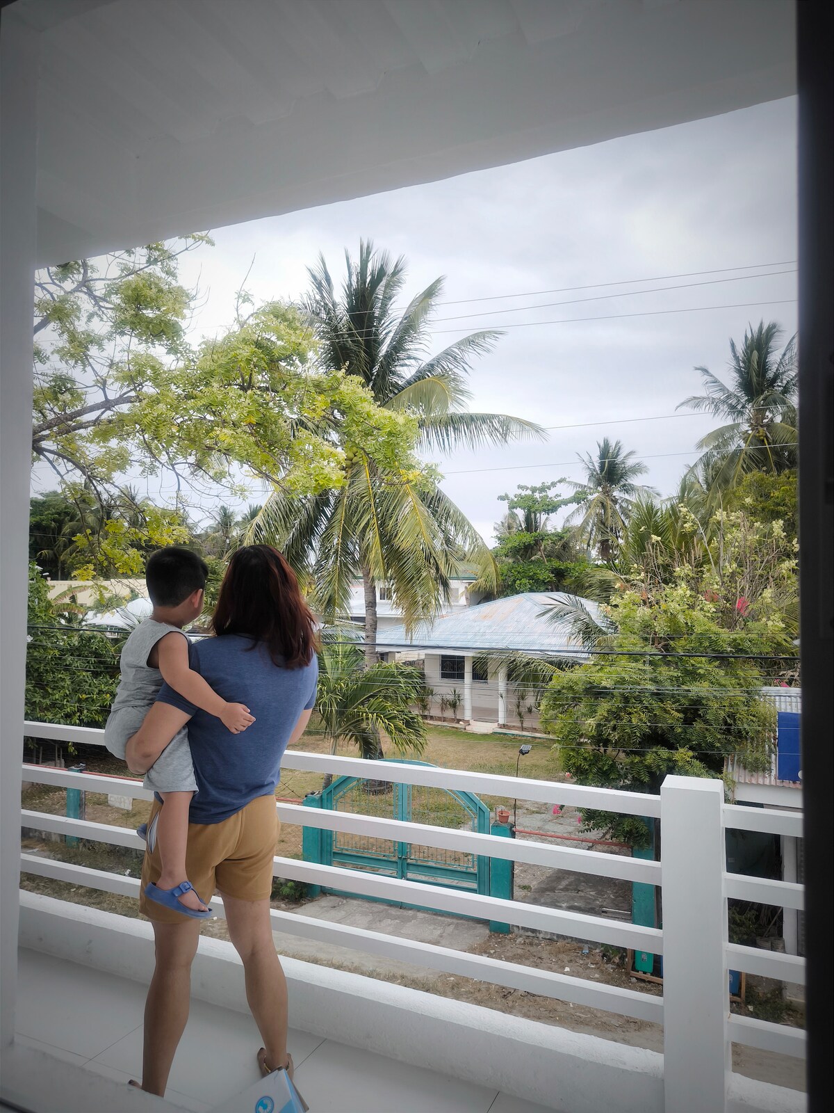 A person is seen holding a child while standing on a balcony. The view features palm trees and greenery, with houses visible in the background. The atmosphere conveys a sense of connection to the outdoors.