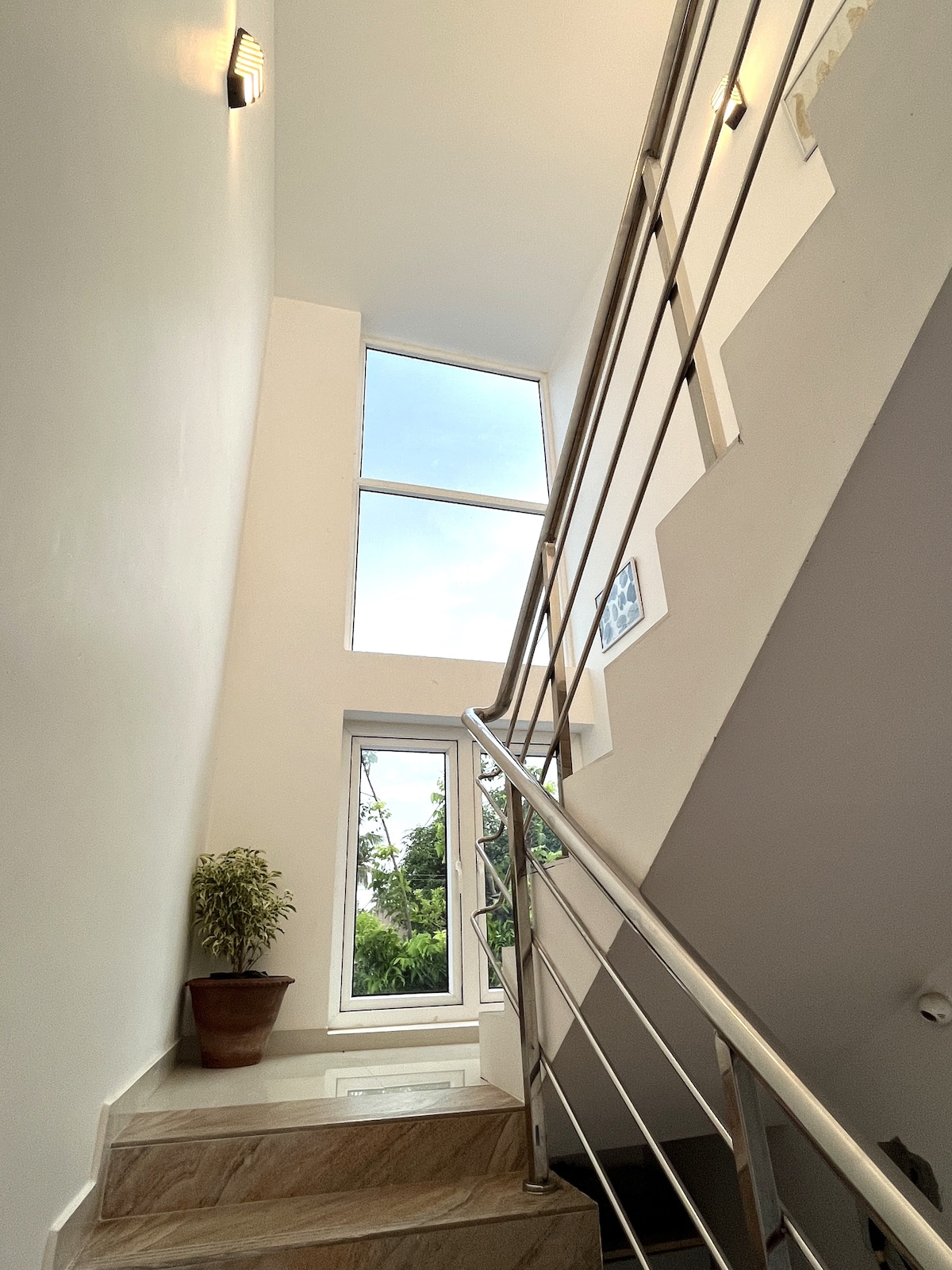 A well-lit staircase is showcased, featuring sleek metal handrails and light-colored walls. Natural light floods in through a large window, illuminating the stairway. A potted plant is positioned near the base of the stairs, adding a touch of greenery to the space.