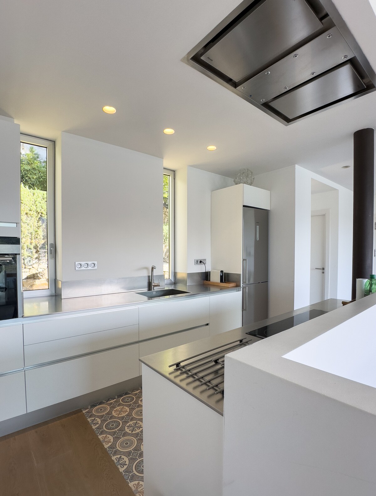 A modern kitchen features sleek white cabinetry and a spacious countertop. Natural light is welcomed through large windows. Stainless steel appliances, including a refrigerator and oven, complement the design. The kitchen island includes a built-in cooktop, showcasing a contemporary layout.