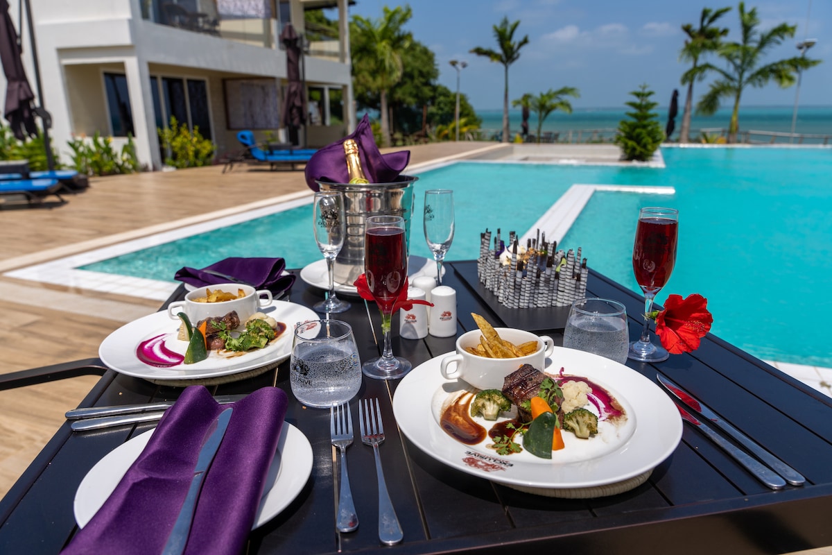 A dining table is set with elegant dinnerware, featuring beautifully plated dishes beside glasses of sparkling beverages. The ocean can be seen in the background, while the pool reflects the sky, creating a serene outdoor dining experience.