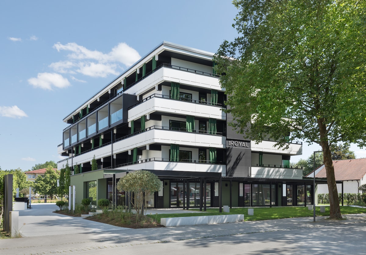The modern building features multiple stories with large glass windows framed by green accents. The landscape is well-maintained, with trees and shrubs surrounding the entrance, creating a fresh and inviting appearance. The name 'DAS ROYAL' is prominently displayed on the façade.