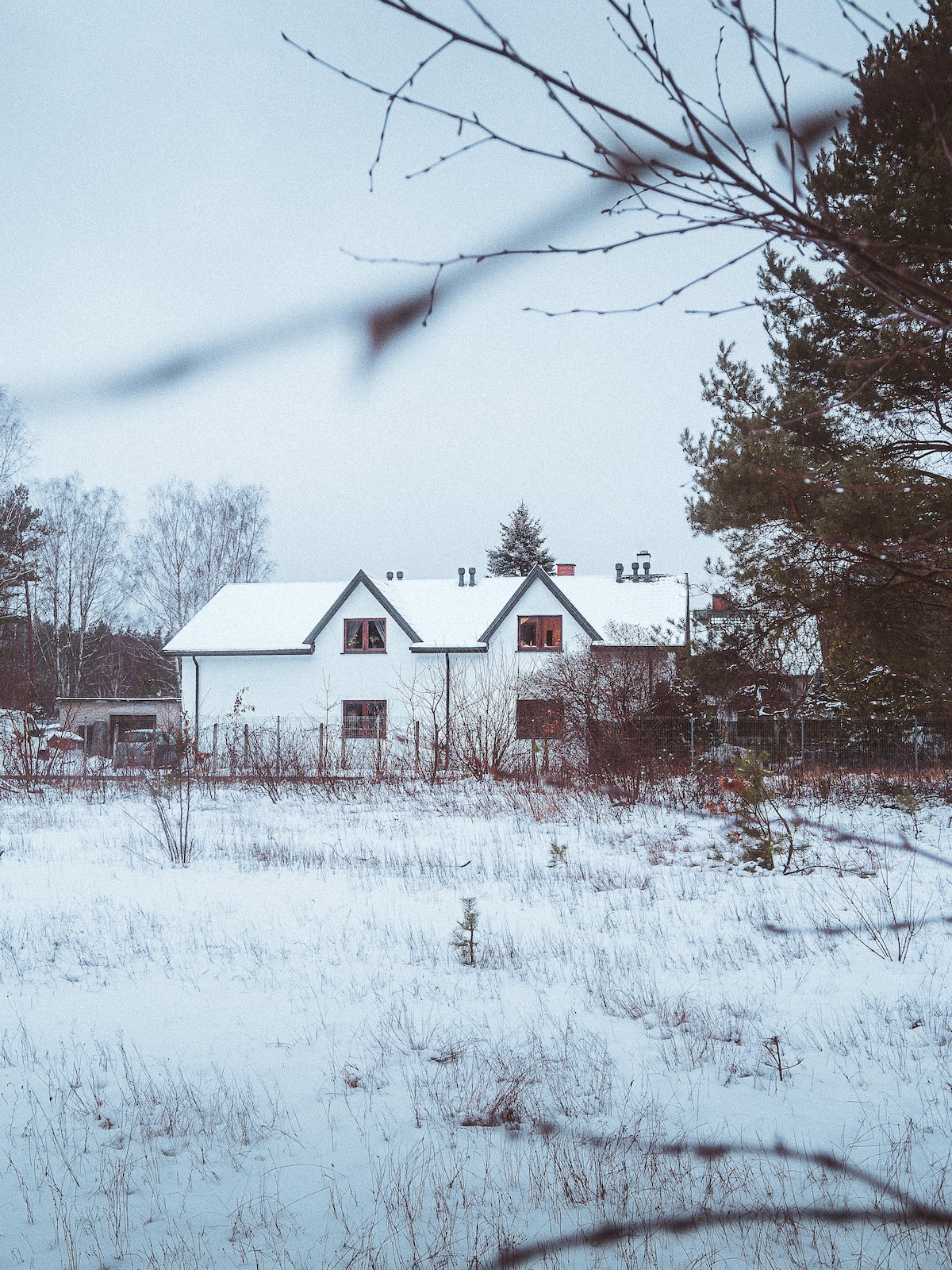 A charming white building is visible in a serene winter landscape, surrounded by a blanket of snow. The architecture features a sloped roof and multiple windows, some of which have warm light glowing within. Leafless trees frame the view, enhancing the quiet atmosphere.