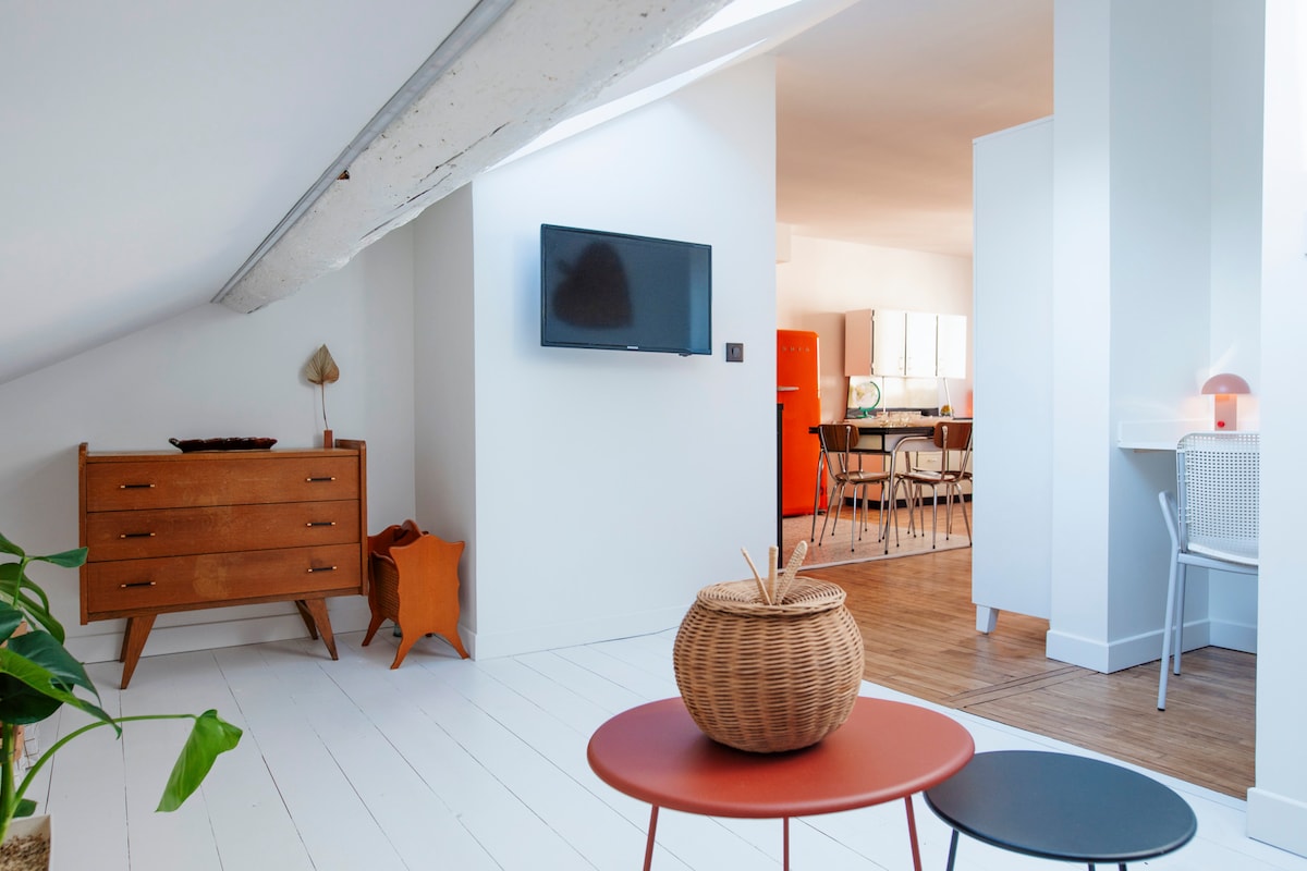 The open layout highlights light-colored wooden floors and a minimalistic design. A wooden dresser and vibrant red refrigerator can be seen in the background, along with a seating area featuring two small tables. Natural light floods the space through the slanted ceiling.