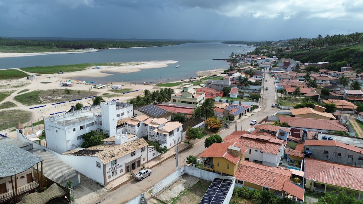 An aerial view captures a coastal village with a sandy beach and calm waters. The landscape includes a mix of residential buildings, some featuring red roofs, and lush greenery in the background. A riverfront promenade is visible, inviting exploration of the area.