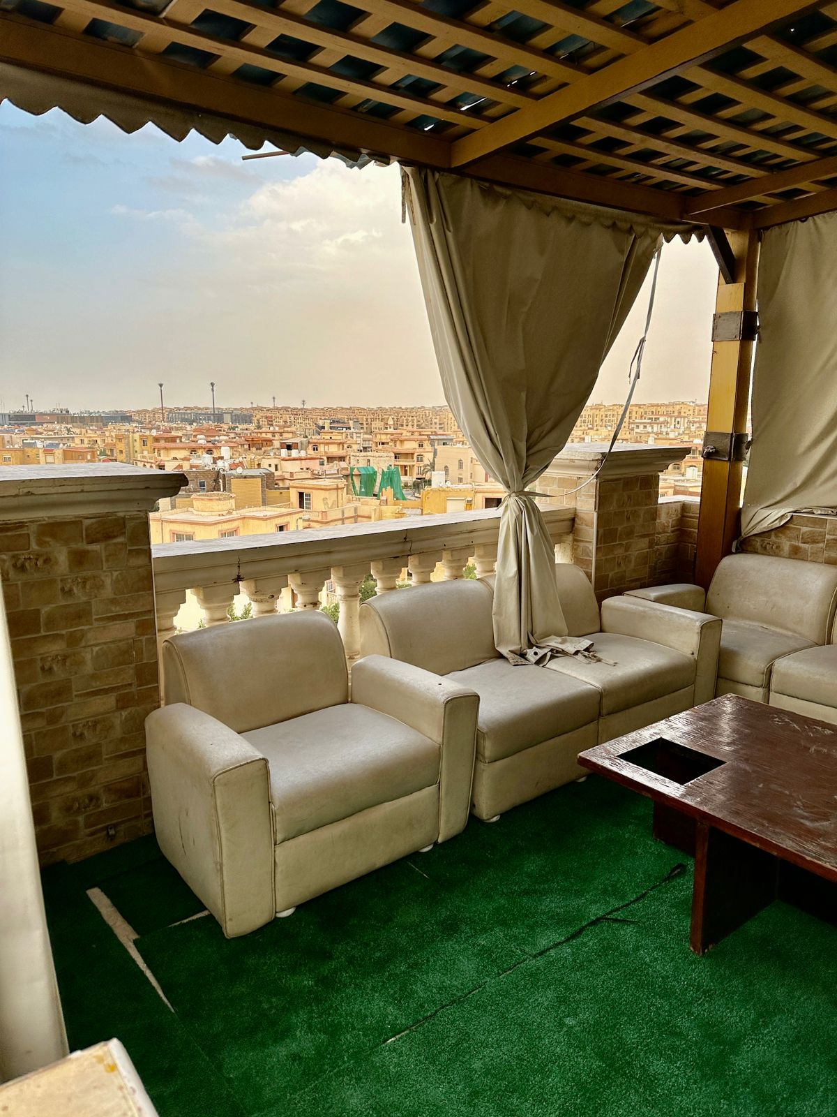 An outdoor terrace is furnished with white upholstered chairs surrounding a wooden table. Soft, green carpet covers the floor, while elegant curtains frame the space, providing privacy. In the distance, a view of rooftops is visible under a partially cloudy sky.