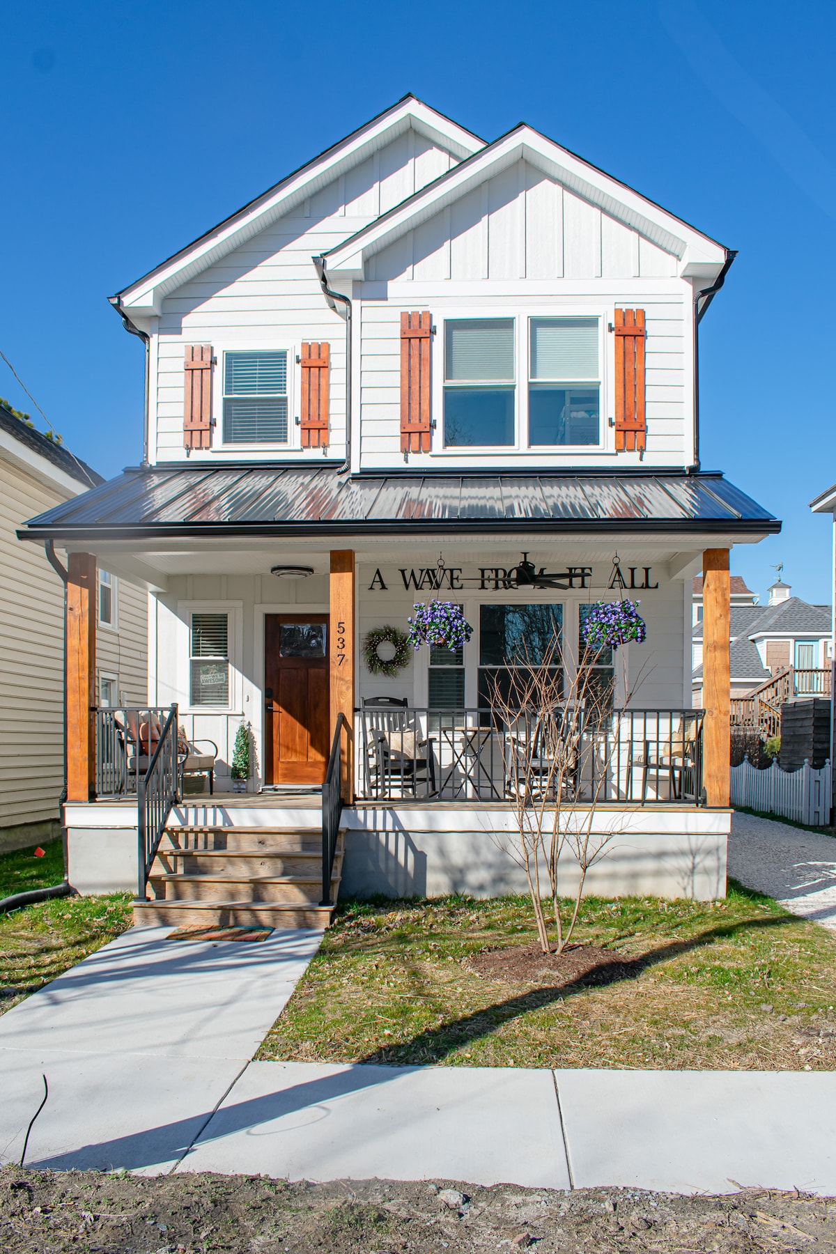 The exterior of the vacation home displays a charming white facade with wooden shutters and a welcoming porch. Greenery frames the entrance, and a pathway leads to the front door, providing easy access. The bright blue sky overhead adds a refreshing atmosphere.