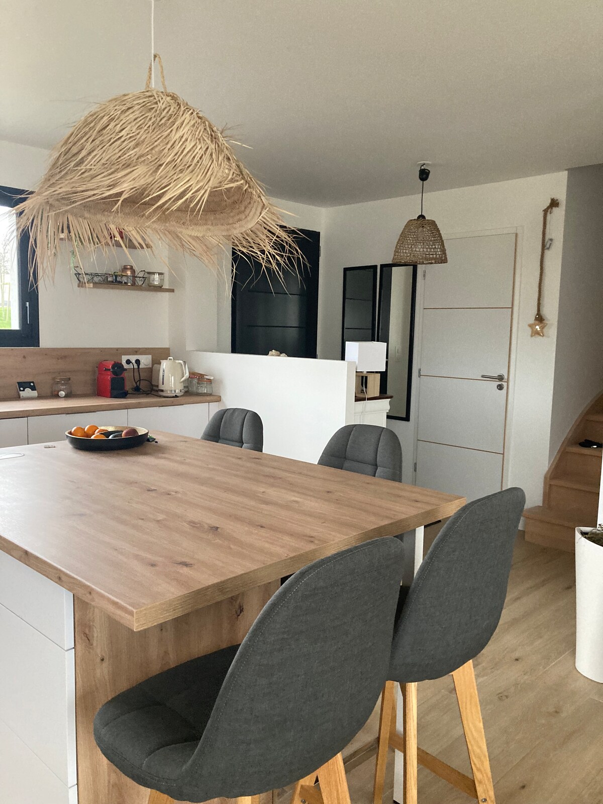 A modern kitchen is displayed, featuring a wooden island with gray bar stools arranged around it. A pendant light made of woven material hangs above. In the background, shelves hold kitchen essentials, and a small dining area complements the space.